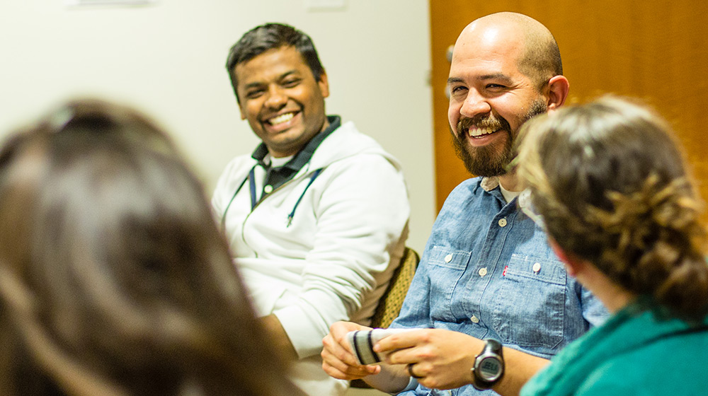 A group of four people sitting and chatting in a room. Two men in focus are smiling and engaged in conversation, with one wearing a white hoodie and the other a blue shirt. The scene appears friendly and relaxed.